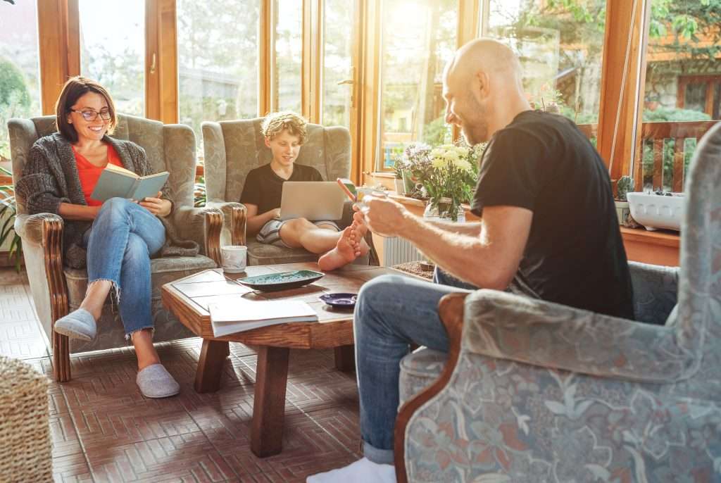 mother, father and son family sitting together at sunroom in cozy armchairs and reading books, using laptop or browsing a smartphone.